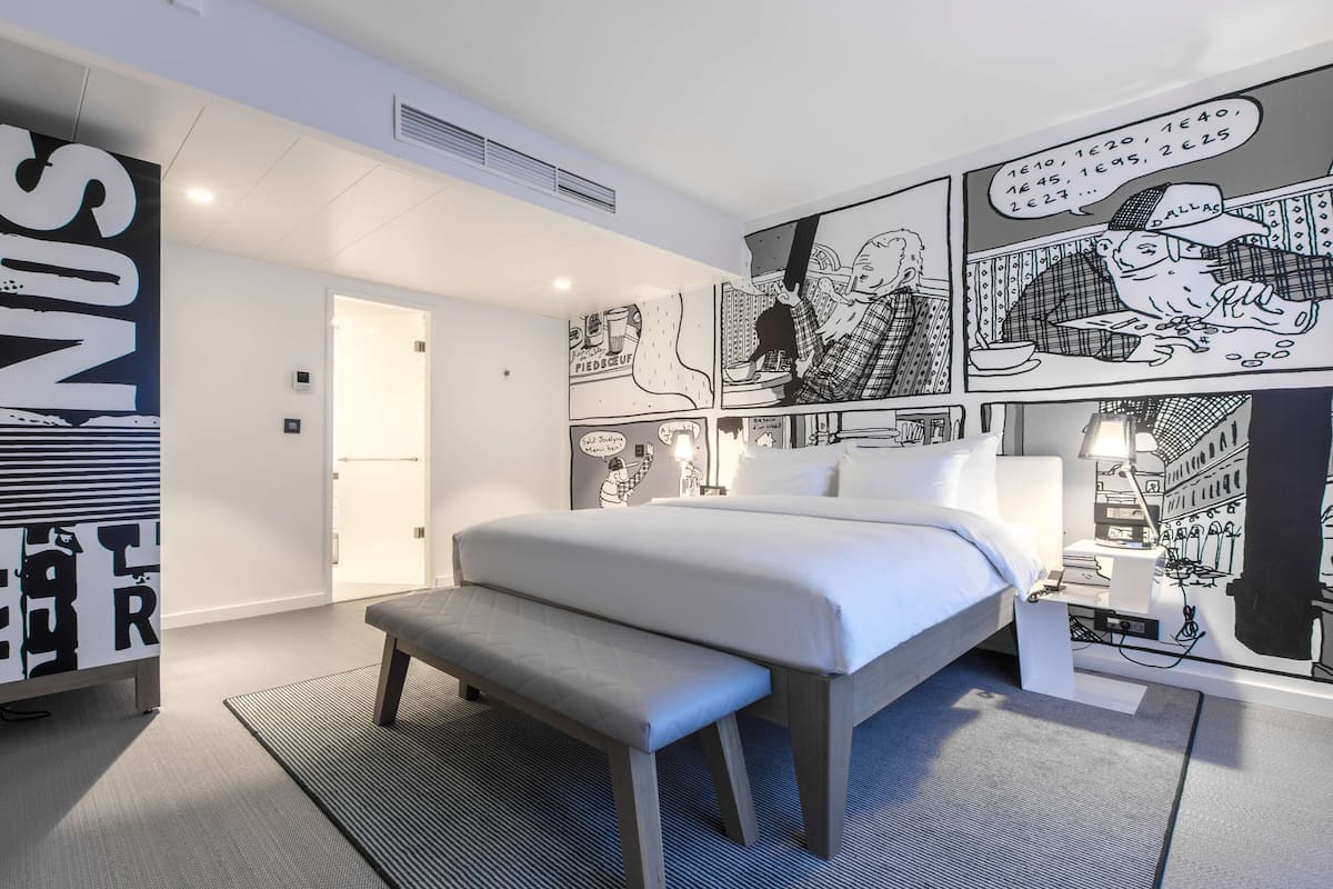 Quarto de hotel moderno com uma cama grande, um banco, uma mesa de cabeceira e uma parede com arte em estilo de história em quadrinhos.