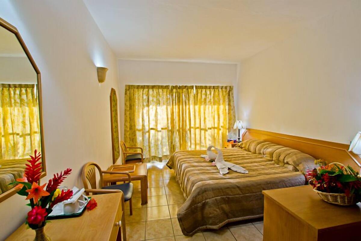 Habitación de hotel con cama, escritorio, silla, espejo y ventana con cortinas.