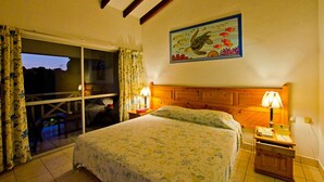 Ein Schlafzimmer mit einem Bett, zwei Nachttischen mit Lampen, einem hölzernen Kopfteil, einem großen Fenster mit Vorhängen und einem Gemälde einer Schildkröte an der Wand.