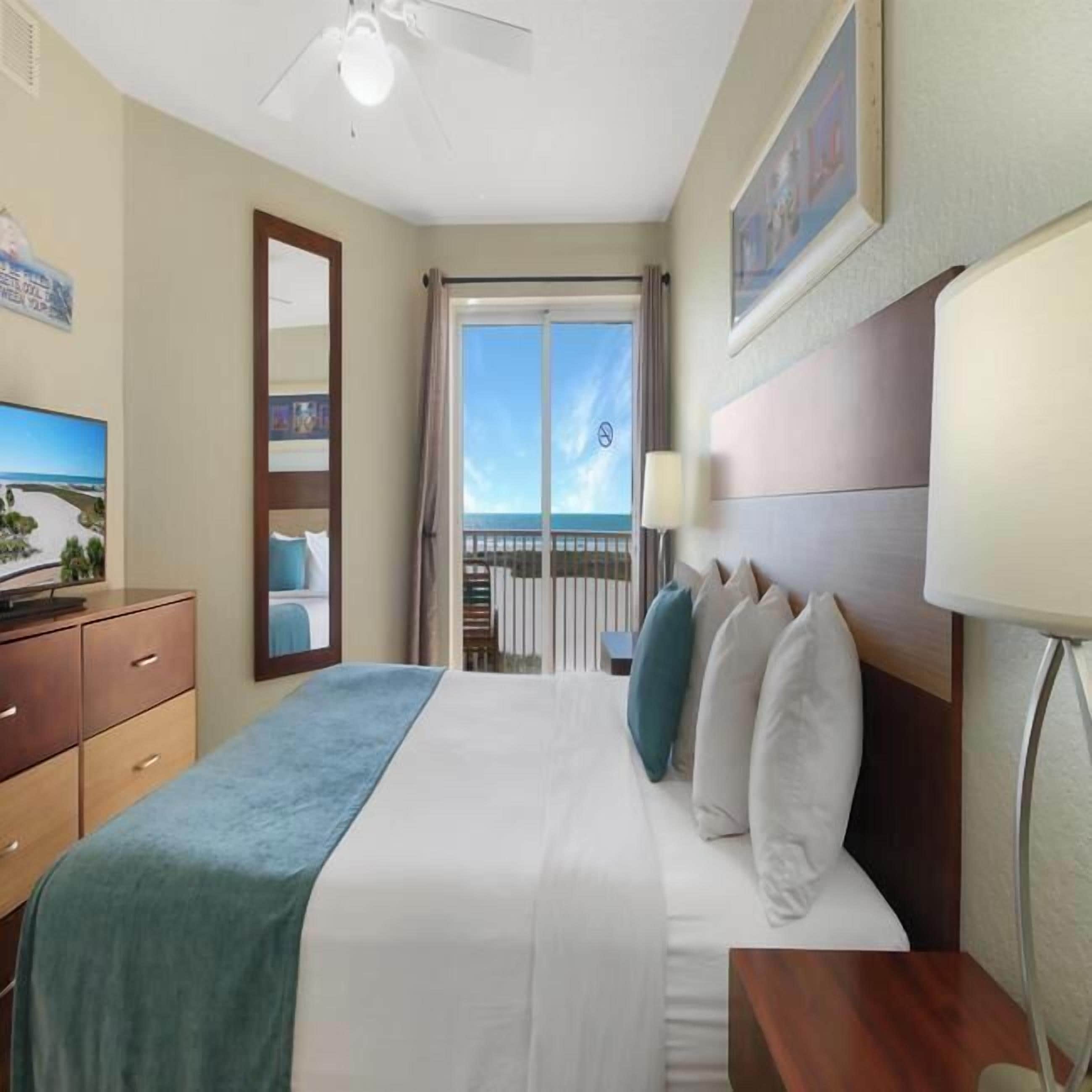 A hotel room with a bed, a desk, a dresser, a ceiling fan, and a balcony with a view of the sea.