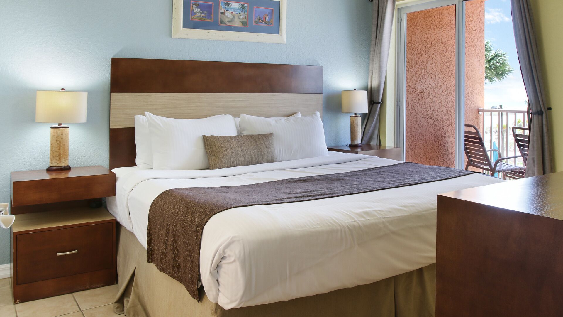 Habitación de hotel con una cama grande, dos mesitas de noche, un cabecero de madera y un balcón con sillas.
