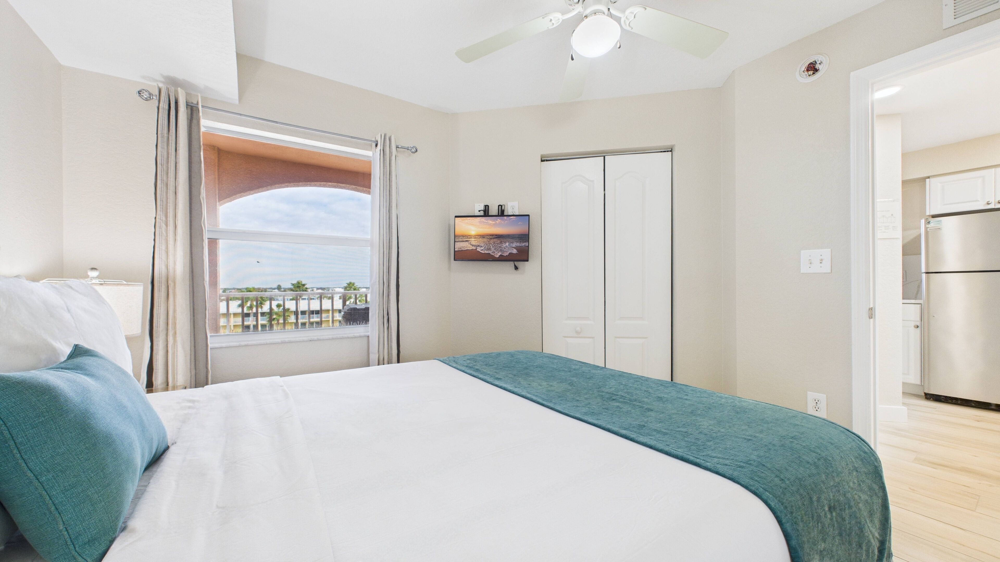 A bedroom with a bed, a view of the sea, a ceiling fan, and a kitchen area.
