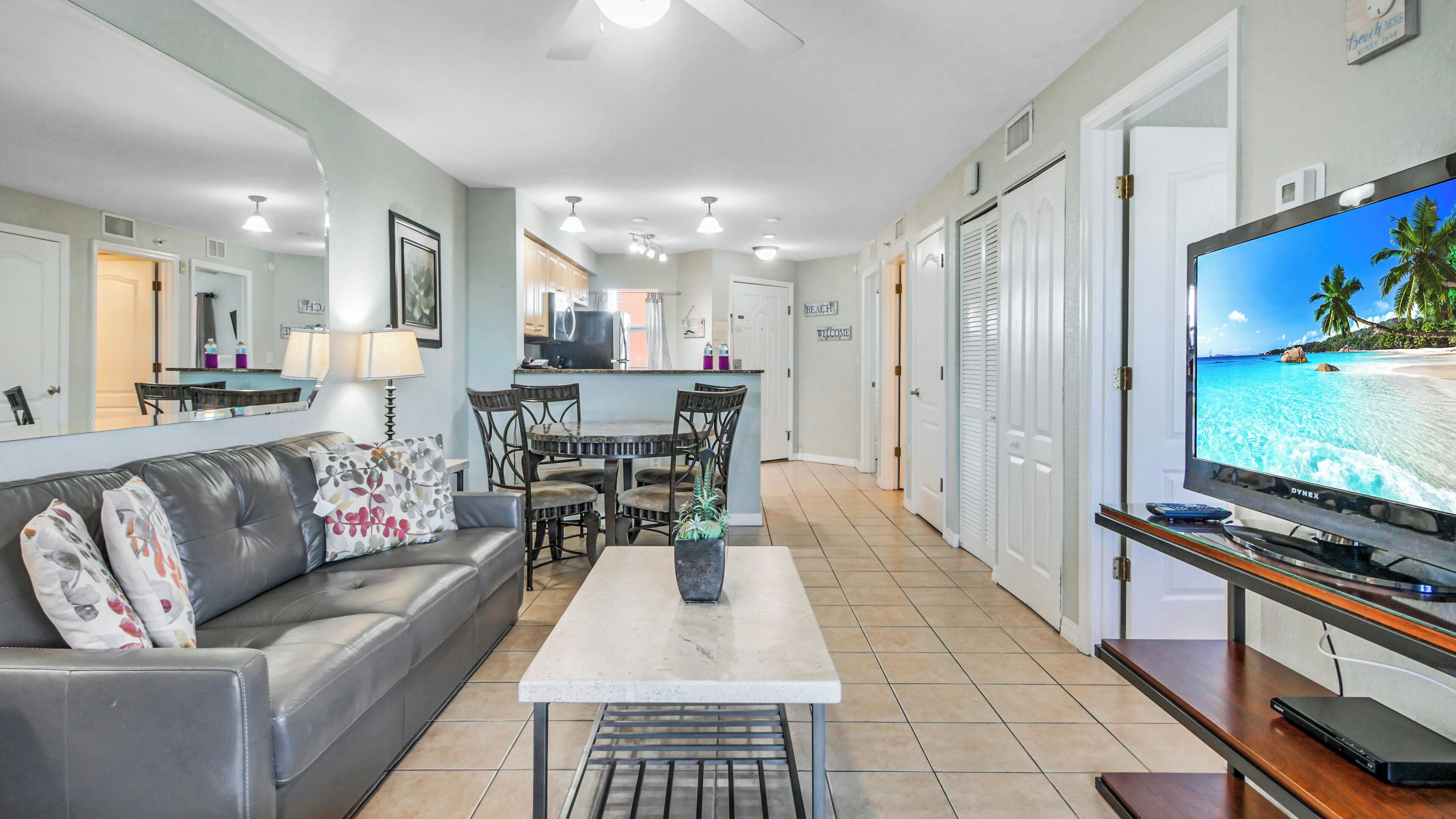 Un salon moderne avec un canapé gris, une table basse et un téléviseur diffusant une scène de plage. On aperçoit, au fond, un coin cuisine avec une table à manger et des chaises.