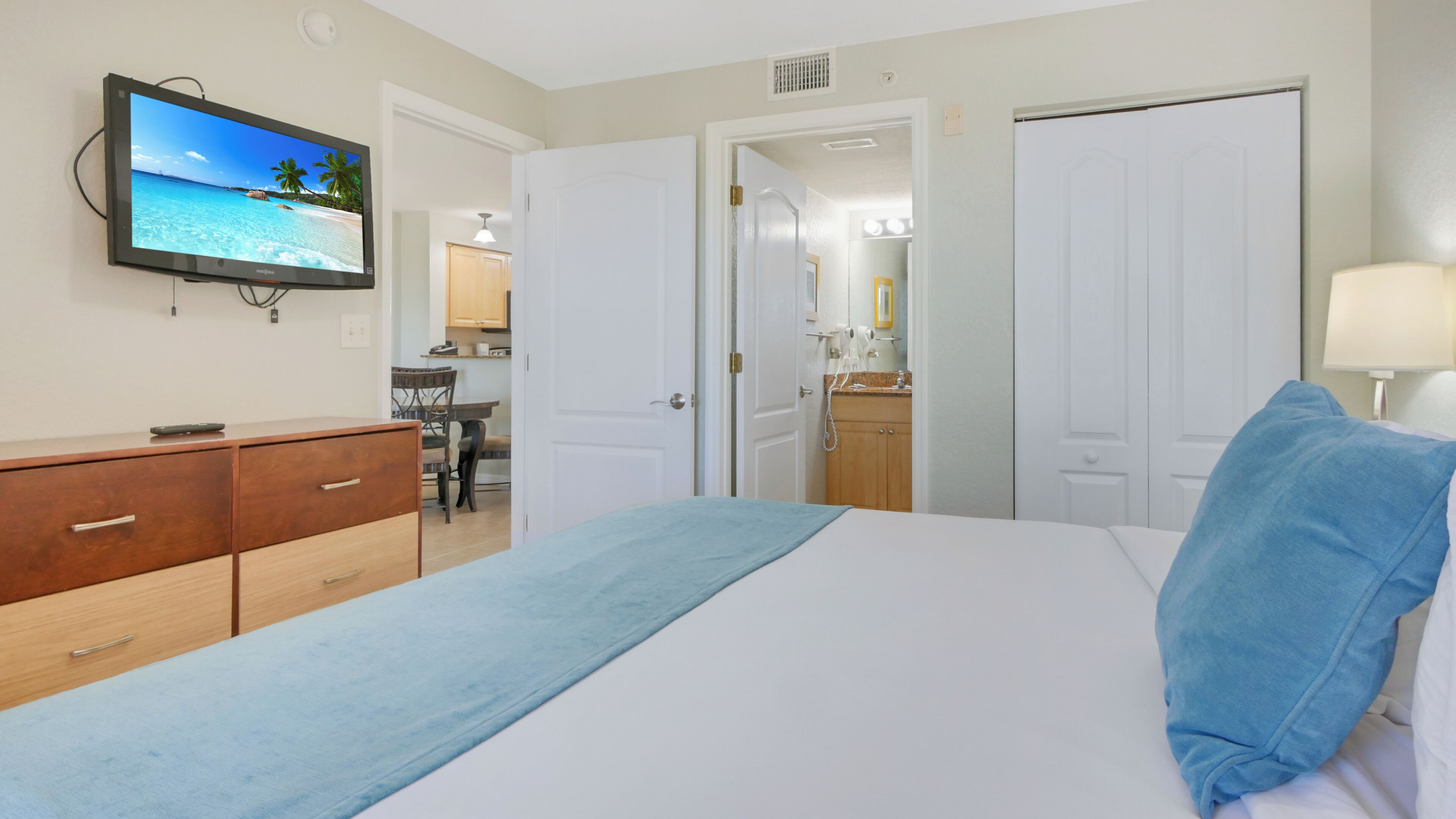 Ein Hotelzimmer mit Bett, Fernseher zeigt eine Strandansicht, Schreibtisch mit Stuhl und einem durchgegangenen Tür sichtbaren Badezimmer.