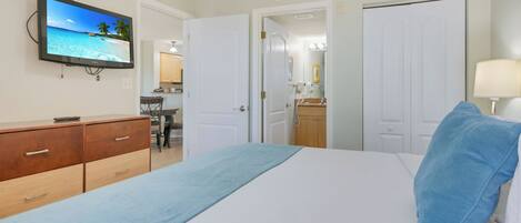 Ein Hotelzimmer mit Bett, Fernseher zeigt eine Strandansicht, Schreibtisch mit Stuhl und einem durchgegangenen Tür sichtbaren Badezimmer.