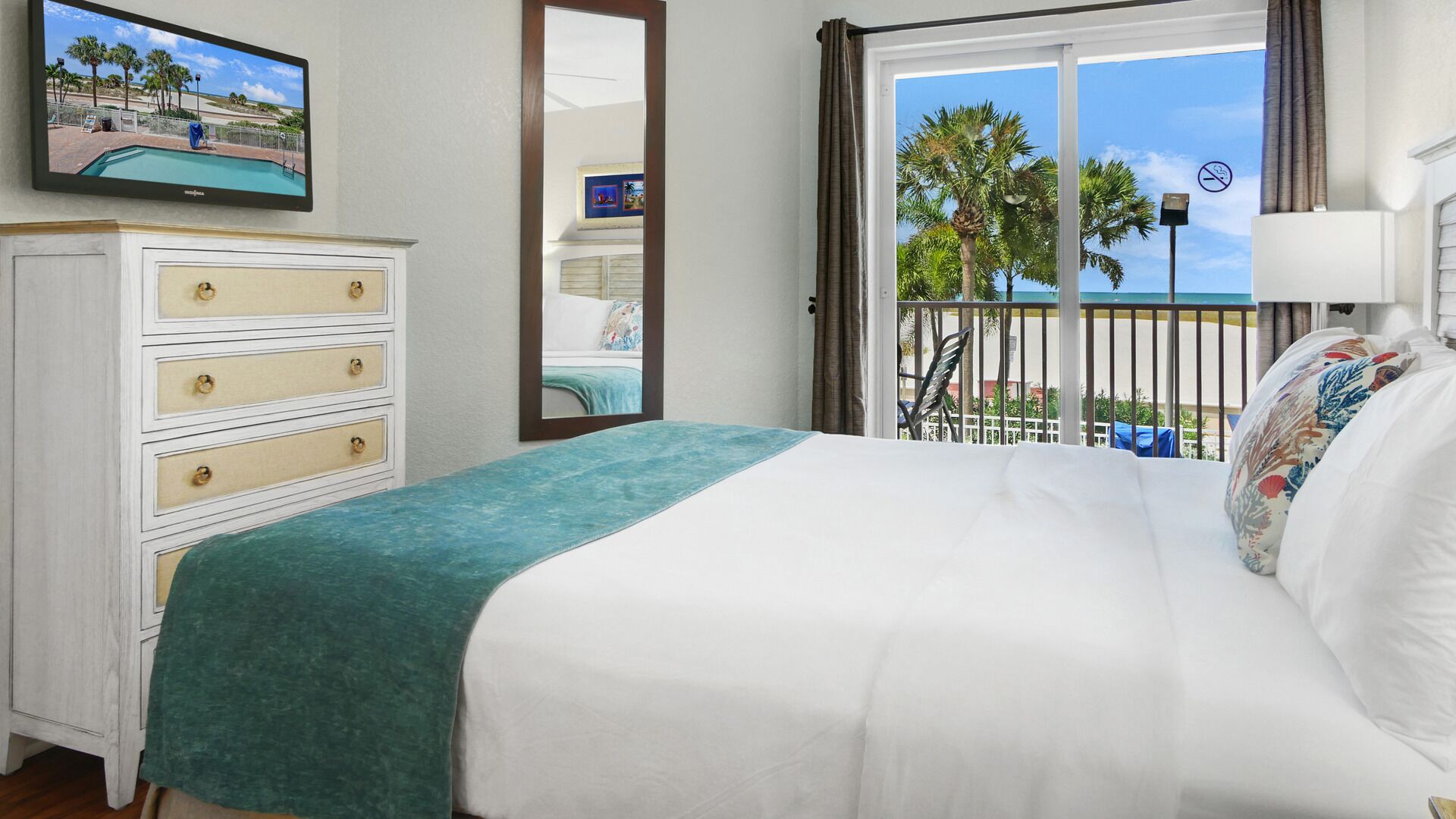 A hotel room with a bed, a dresser, a TV, a balcony with a view of palm trees and a beach, and a window with a view of the beach.