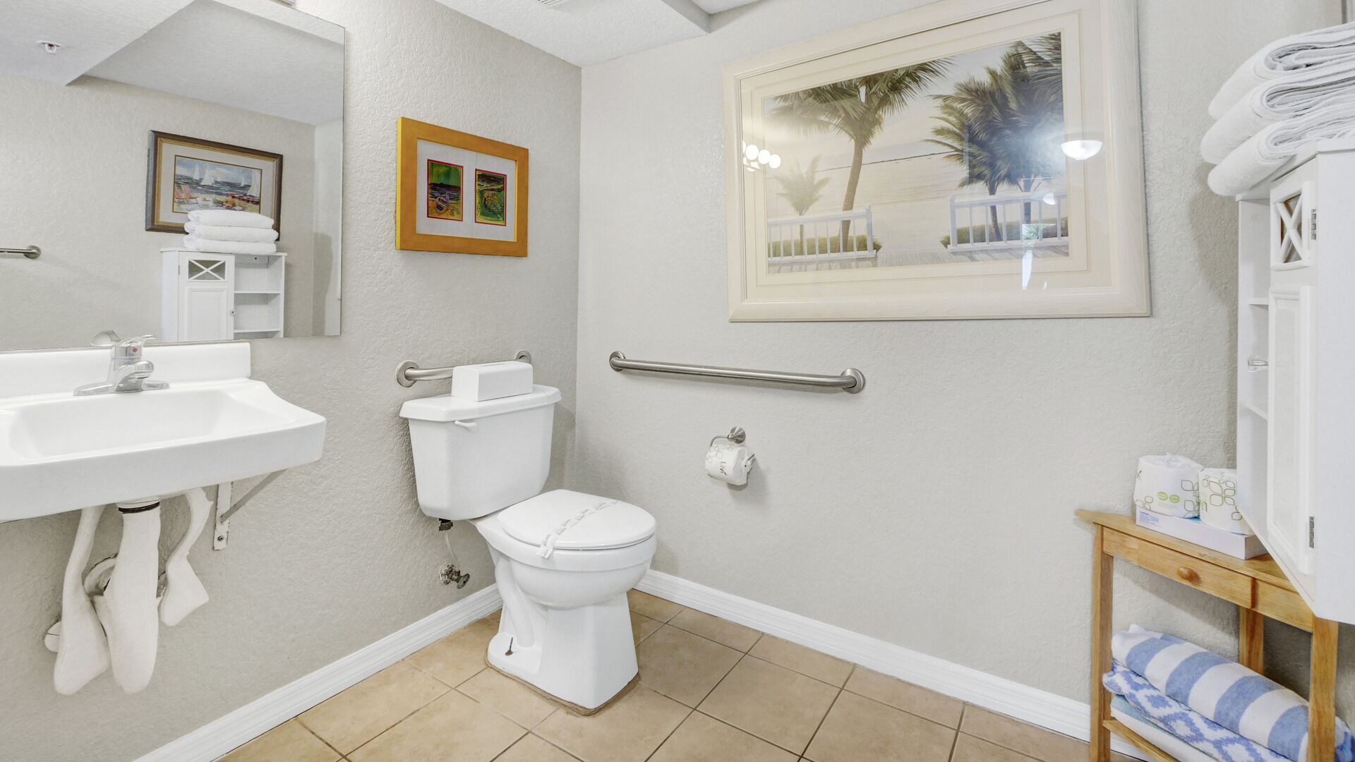 A bathroom with a sink, toilet, and a handrail. There is a framed picture on the wall and a towel rack with towels.
