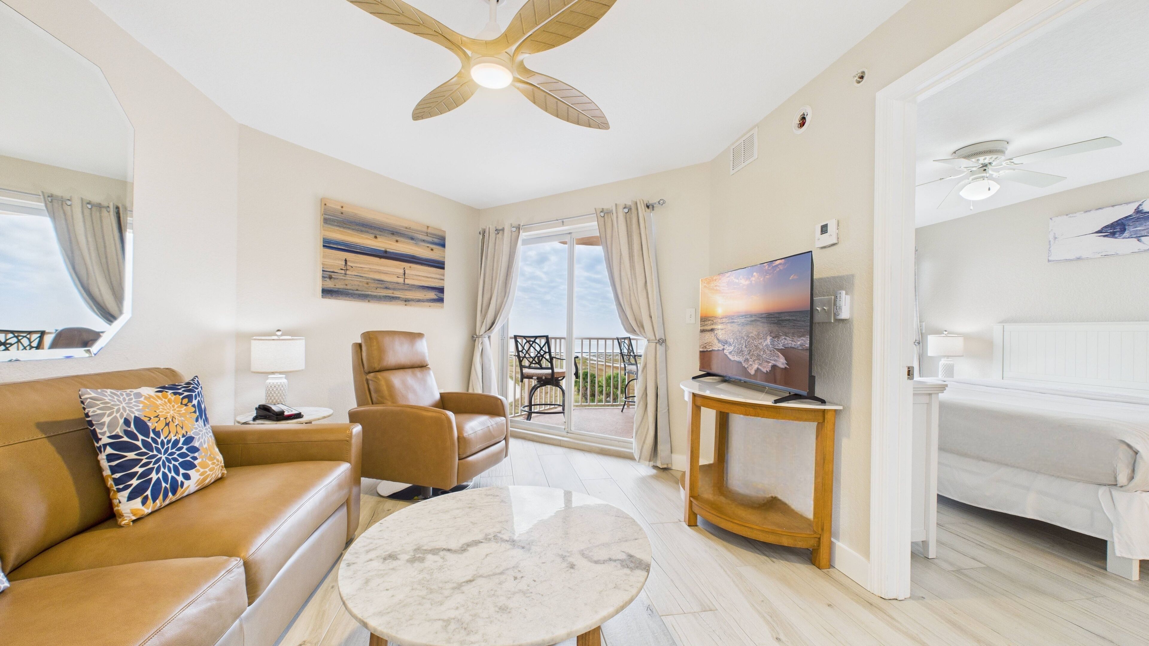 A spacious living room with a sofa, armchair, and a TV on a stand. There is a balcony with a view of the ocean.
