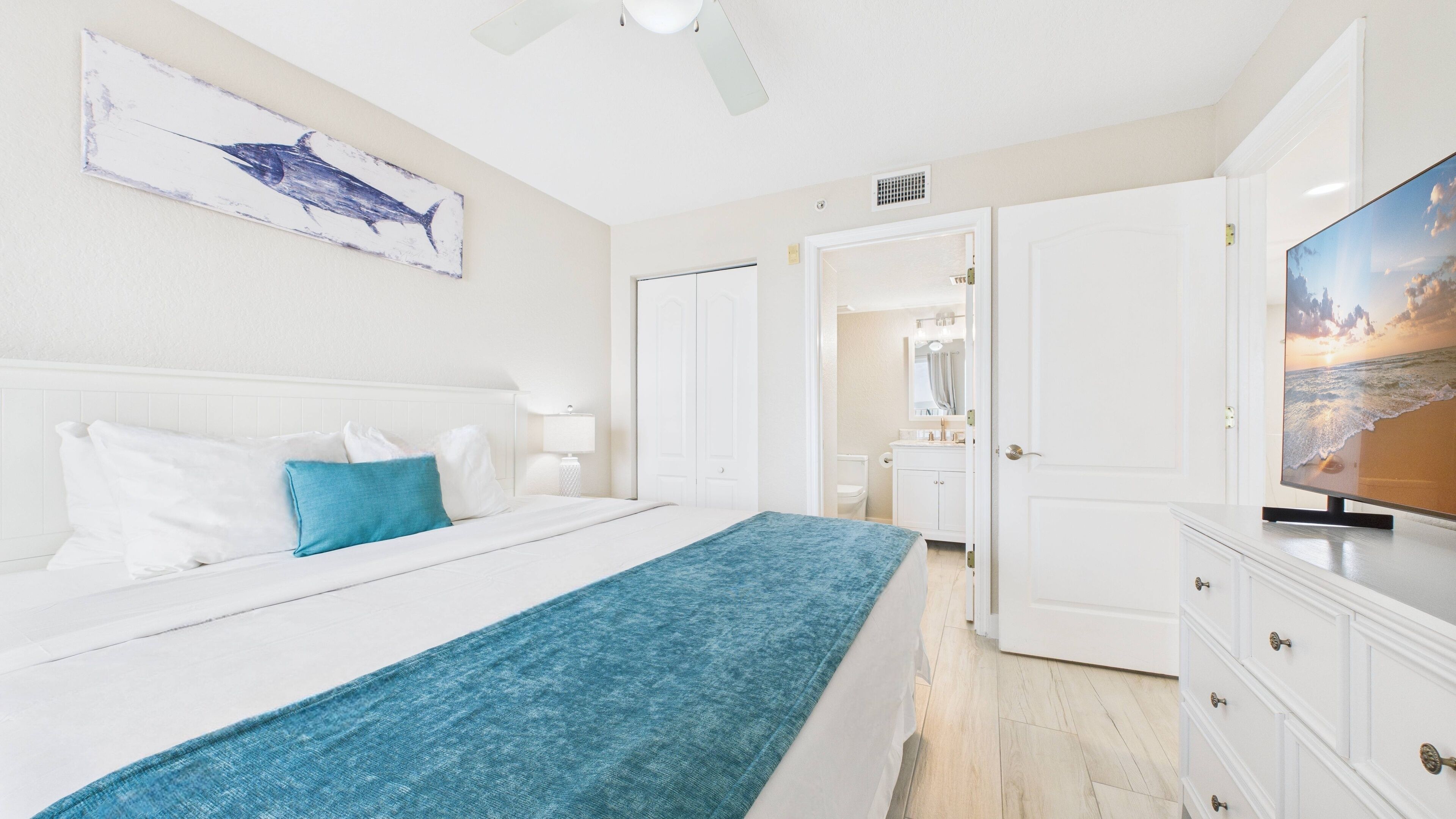 A bedroom with a bed, a blue throw blanket, a white headboard, a painting of a shark, a TV, and a bathroom visible through an open door.