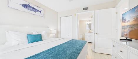 A bedroom with a bed, a blue throw blanket, a white headboard, a painting of a shark, a TV, and a bathroom visible through an open door.