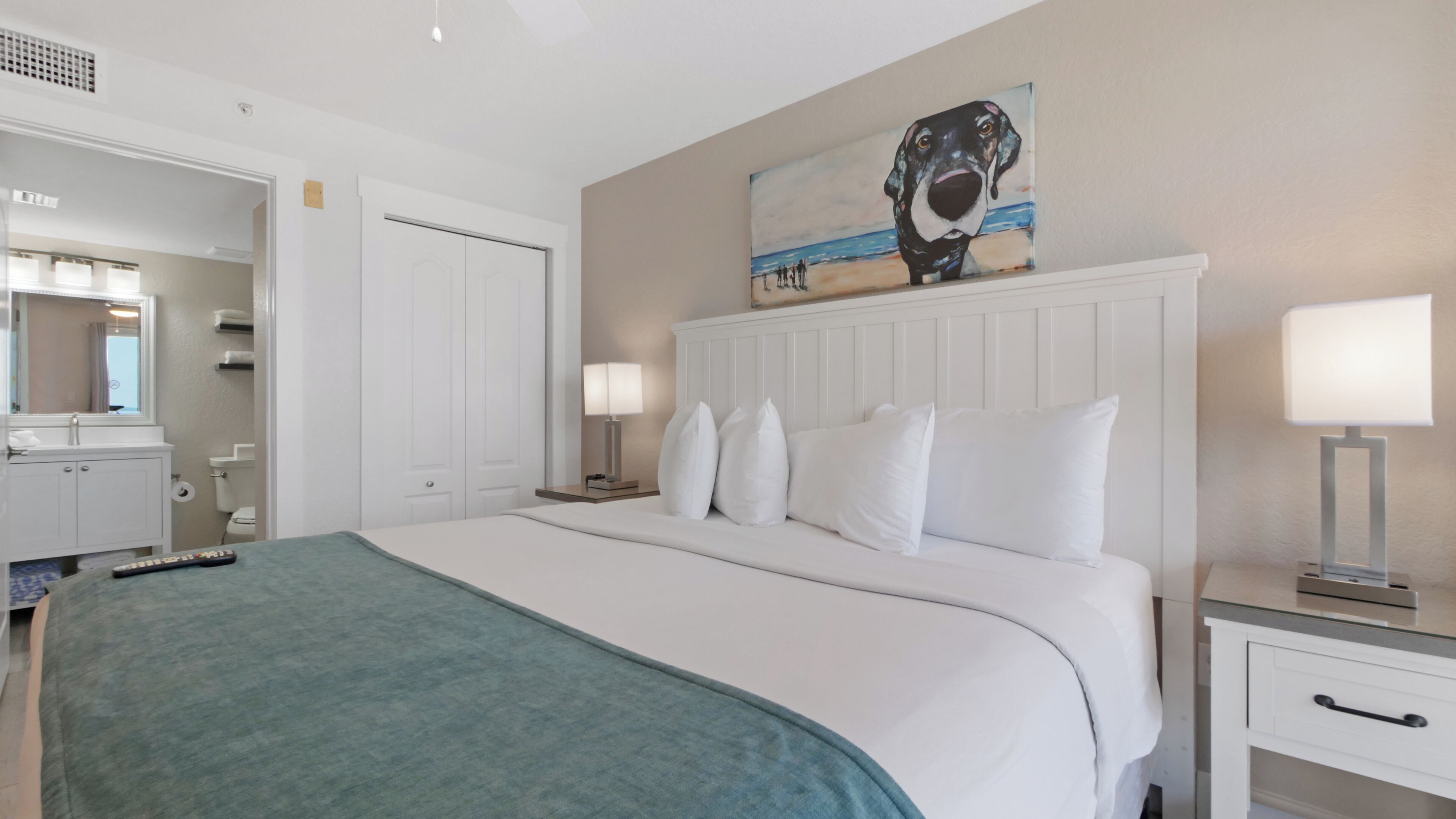 A hotel room with a large bed, white headboard, two bedside tables with lamps, a painting on the wall, and a bathroom visible through an open door.