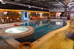 An indoor swimming pool with a hot tub, lounge chairs, and a bar area with seating.