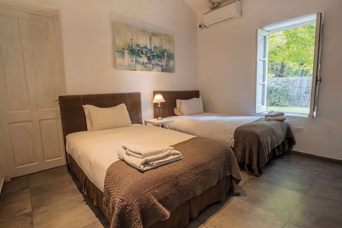 Ein Hotelzimmer mit zwei Betten, einem Bild an der Wand, einem Fenster mit Blick auf Grünflächen und einer Tür.