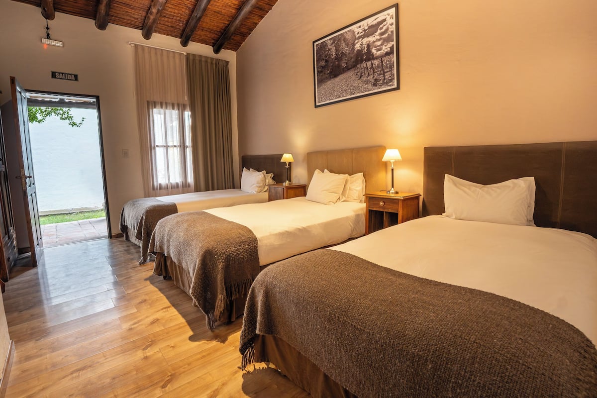 Ein Hotelzimmer mit drei Betten, Holzboden, einer Tür mit der Aufschrift 'SALIDA' und einem gerahmten Bild an der Wand.