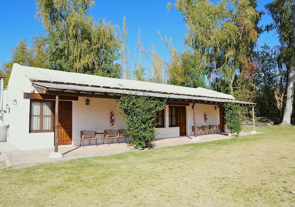 Una edificación de un solo piso con techo plano, un porche cubierto con sillas y un pequeño árbol en la fachada.