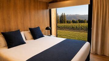 A bedroom with a large bed, two pillows, a bedside lamp, and a view of a vineyard through a window.
