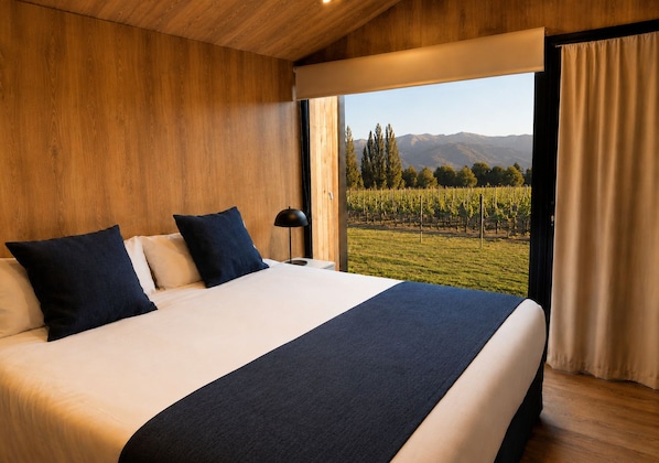 A bedroom with a large bed, two pillows, a bedside lamp, and a view of a vineyard through a window.