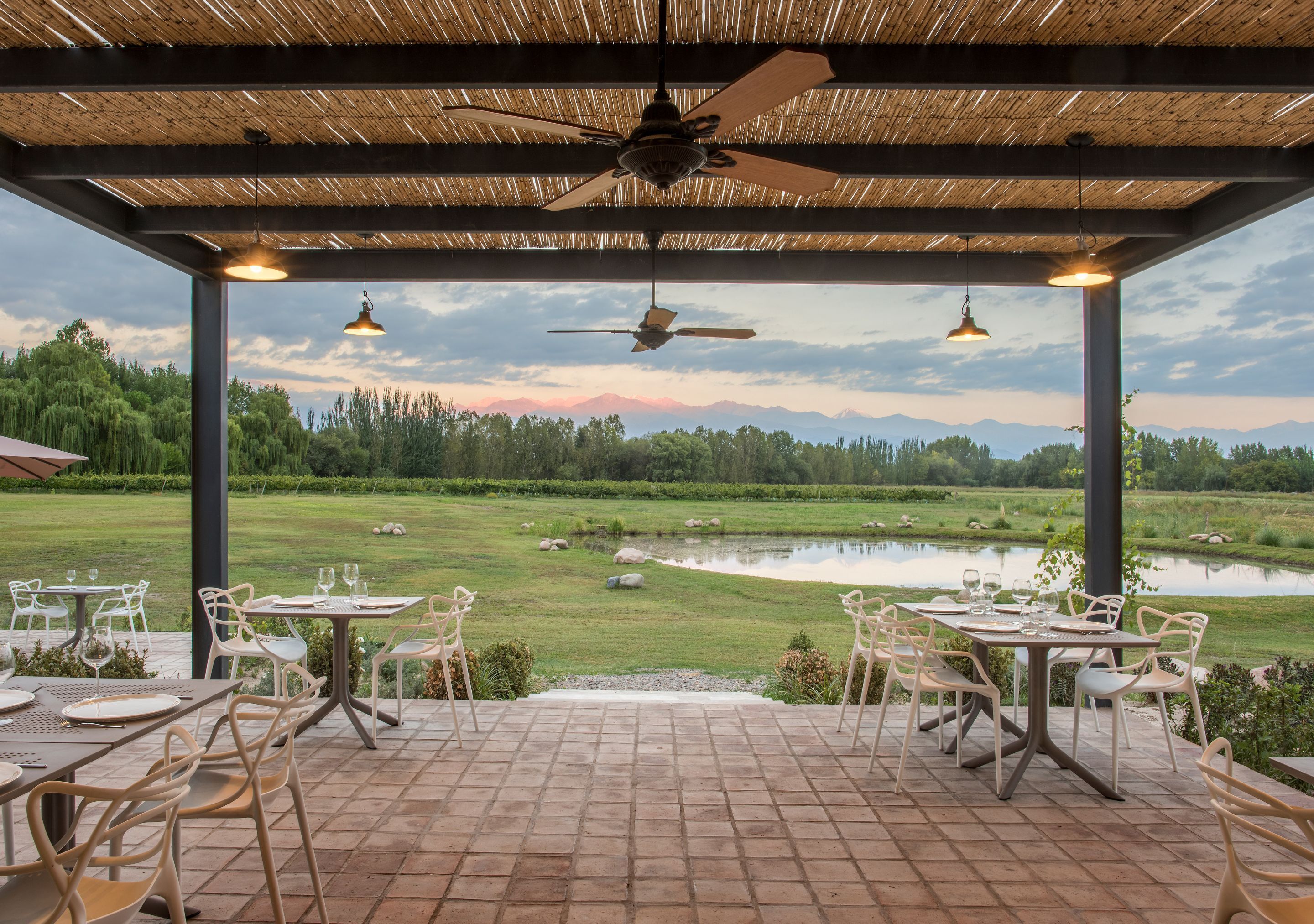 An outdoor patio with tables and chairs, a ceiling fan, and a view of a pond and trees.