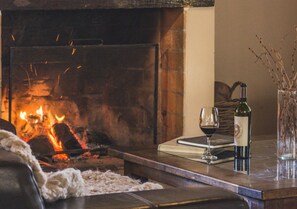 A fireplace with visible flames, a glass of red wine, a bottle of wine, and a vase with branches on a table.