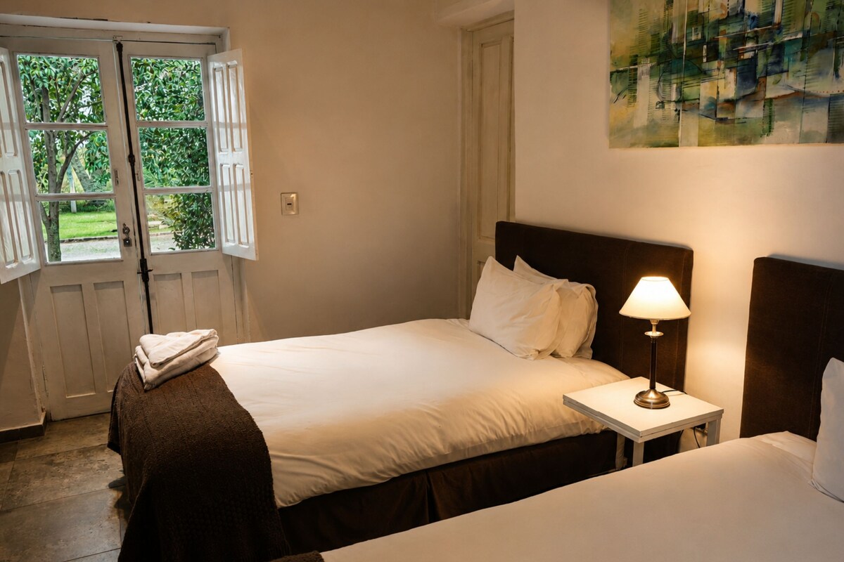 A bedroom with a bed, a nightstand with a lamp, a window with shutters, and a painting on the wall.