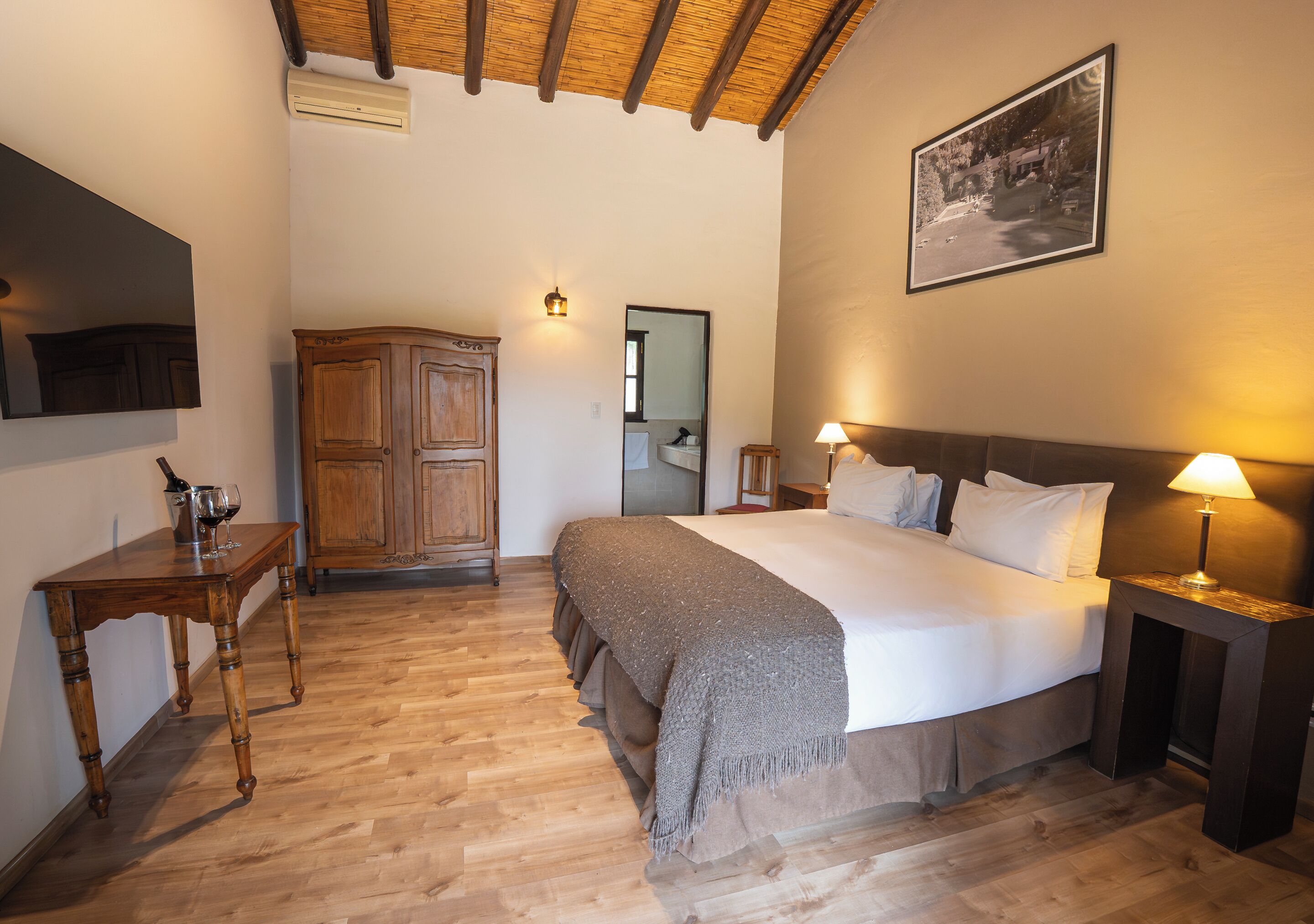 Un dormitorio con piso de madera, una cama con almohadas, una mesita de noche con lámpara, un armario de madera y un cuadro enmarcado en la pared.