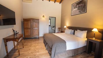 Un dormitorio con piso de madera, una cama con almohadas, una mesita de noche con lámpara, un armario de madera y un cuadro enmarcado en la pared.