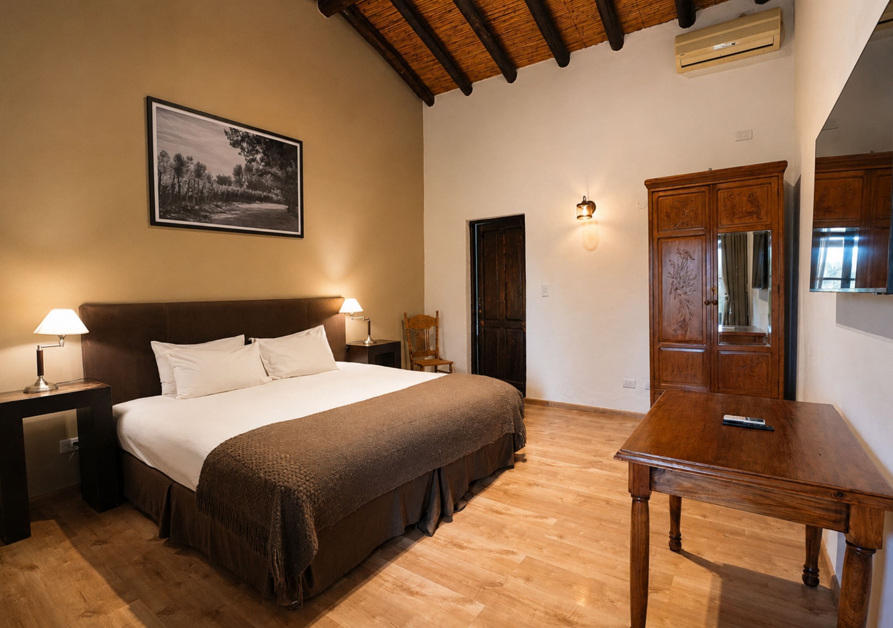 A bedroom with a large bed, wooden nightstands, a wooden table, a wardrobe, and a framed picture on the wall.