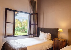 Une chambre d’hôtel avec une grande fenêtre, un lit avec du linge de maison blanc, une table de chevet en bois avec une lampe, et une vue sur la verdure à l’extérieur.