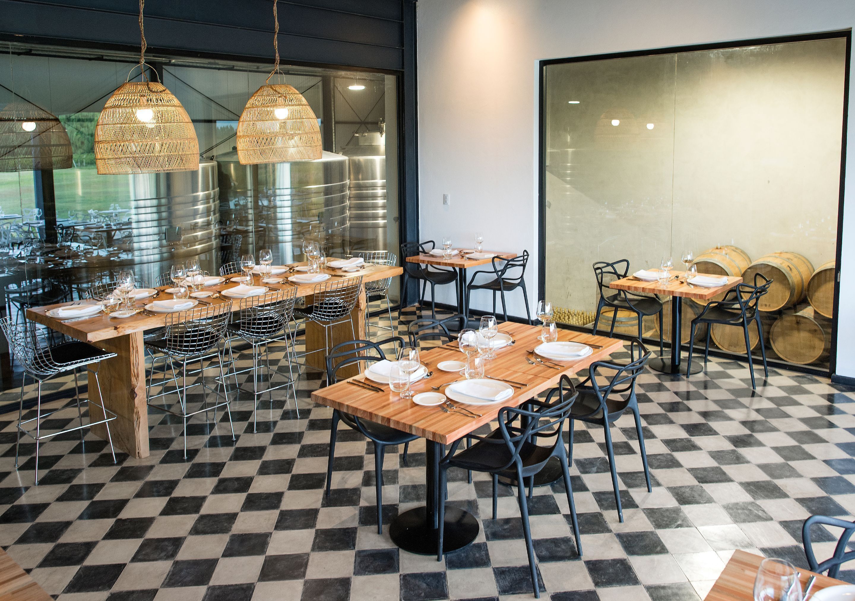 Interior de restaurante con suelo a cuadros, varias mesas preparadas para comer, lámparas de mimbre colgantes y tanques de acero inoxidable al fondo.