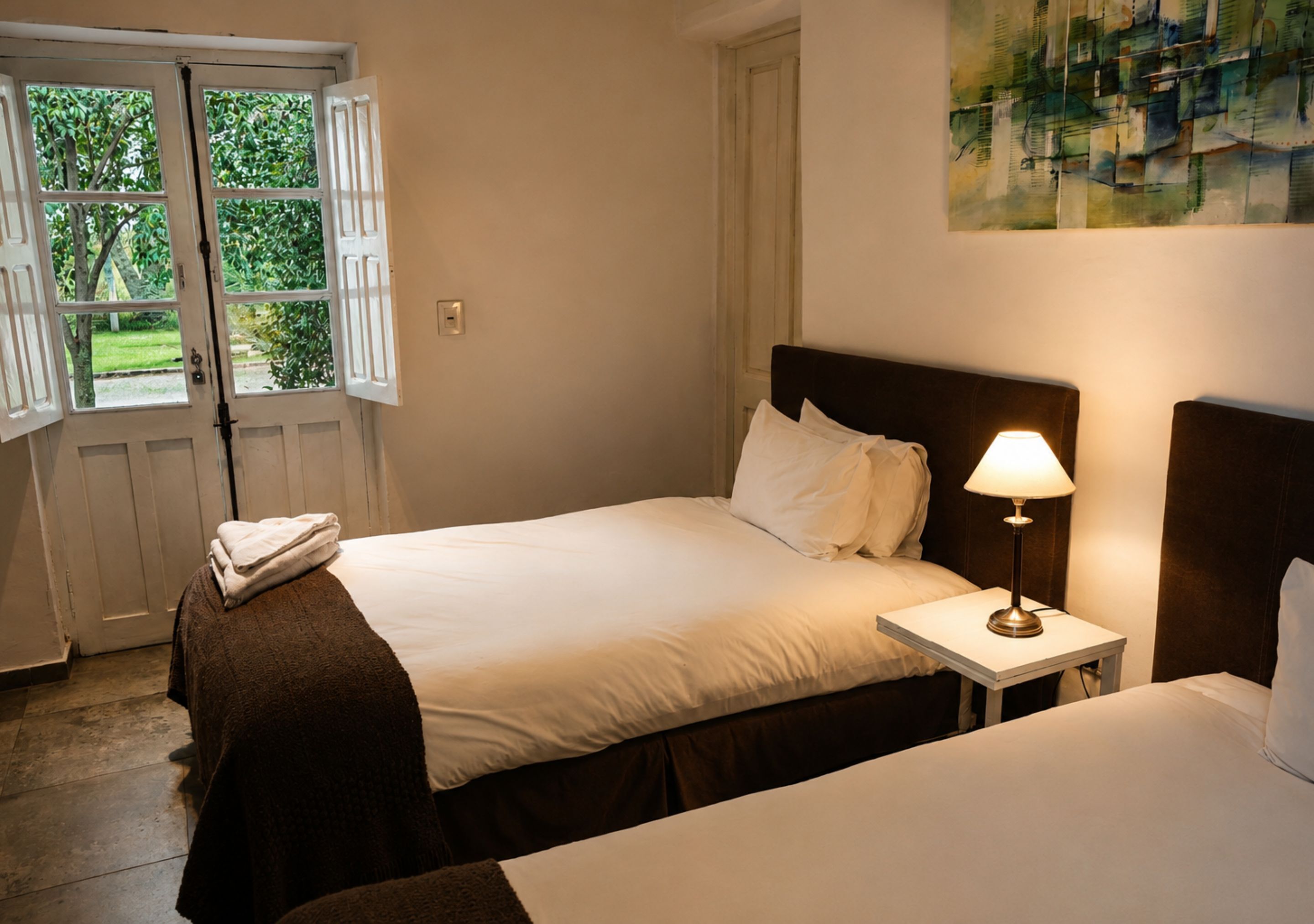 A bedroom with a bed, a nightstand with a lamp, a window with shutters, and a painting on the wall.