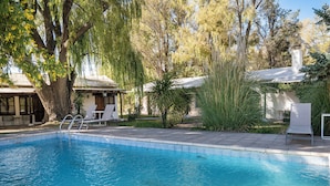 A swimming pool with a tiled edge, surrounded by trees and a building with a covered patio area. - Postales Finca & Bodega by DOT Boutique (Tunuyan)