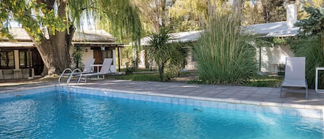 Une piscine dont le bord est revêtu de carreaux, entourée d’arbres et bordée d’un bâtiment doté d’un patio couvert.