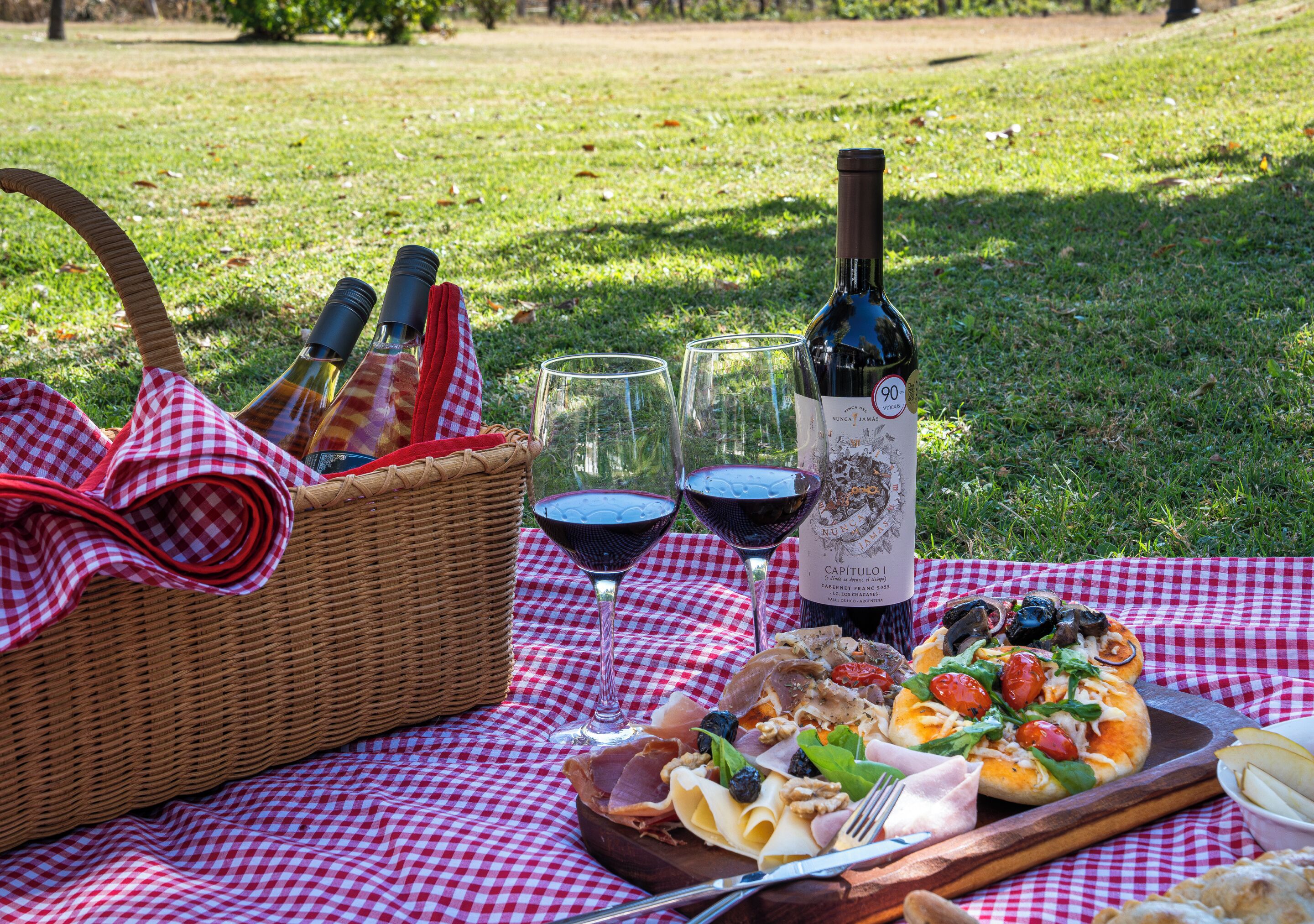 A picnic setup with a basket, wine bottle, glasses, and a pizza on a checkered tablecloth.