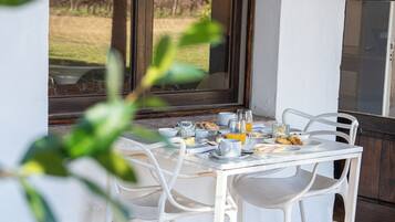 Una mesa de desayuno dispuesta con platos, tazas y un vaso de jugo de naranja, ubicada cerca de una ventana con vistas a un área verde.