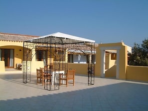 A covered outdoor seating area with a table and chairs, a pergola structure, and a yellow building in the background. - Papillo Hotels & Resorts Borgo Antico (Olbia)