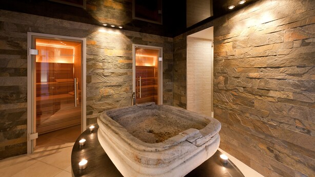 Una bañera de piedra en un área de spa, con dos puertas de cristal y un muro de piedra al fondo.