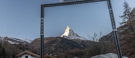 Een berg met een top, een houten chalet en een metalen bord waarop "Welcome" en "hemizeus.ch" staan.