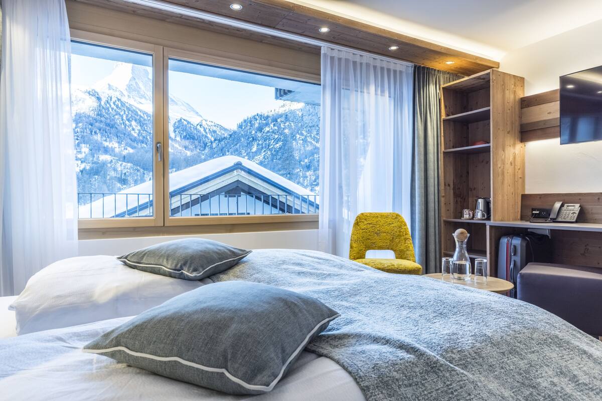 Ett hotellrum med en säng, en stol, en tv och utsikt över snötäckta berg.
