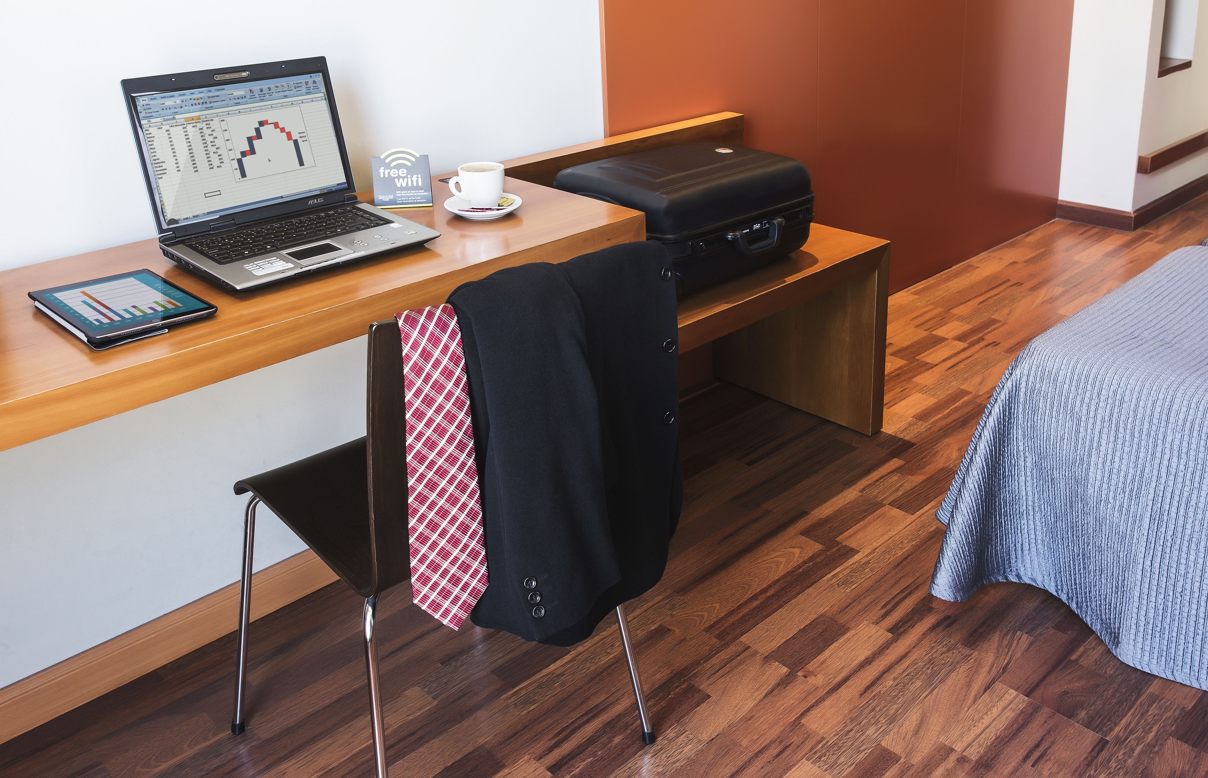 Habitación de hotel con un escritorio de madera, una computadora portátil, un teléfono inteligente, una maleta y una cama con una manta azul.