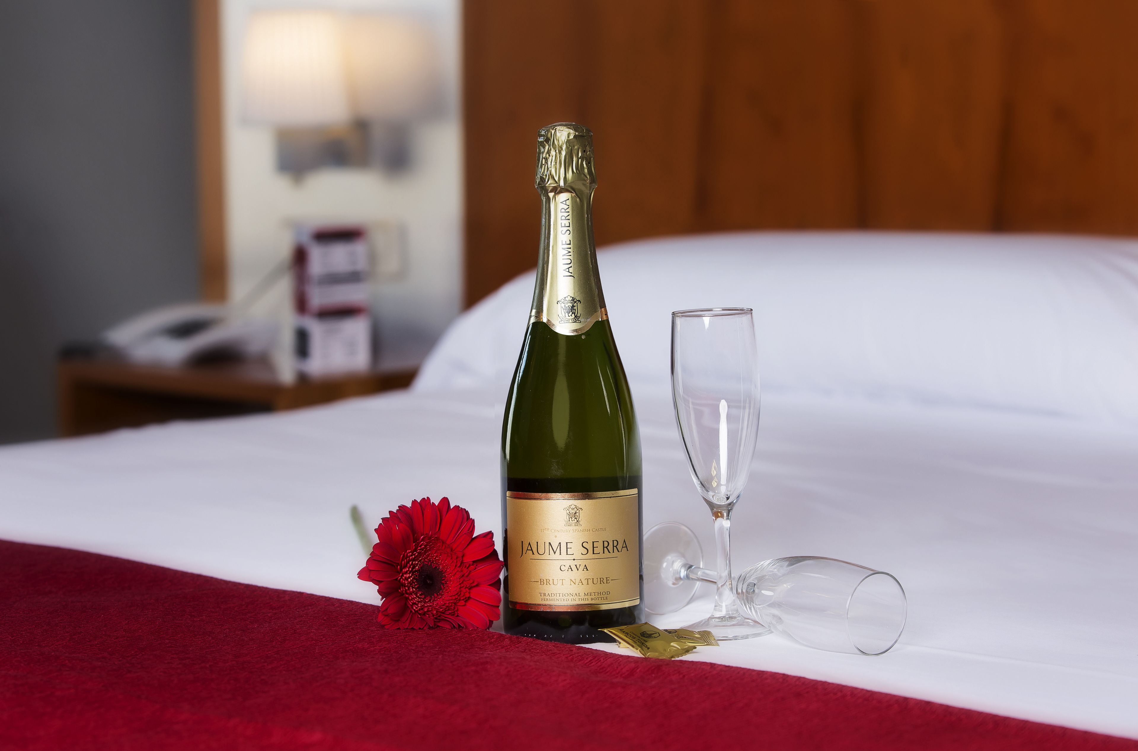 Una botella de cava Jaume Serra, una copa y una flor roja sobre una cama con sábanas blancas.