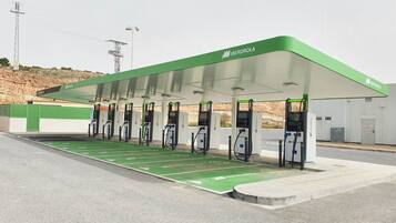 Eine Ladestation für Elektroautos mit grüner Dachbegrünung, mehreren Ladepunkten, einem überdachten Wartebereich und einem gepflasterten Parkplatz.