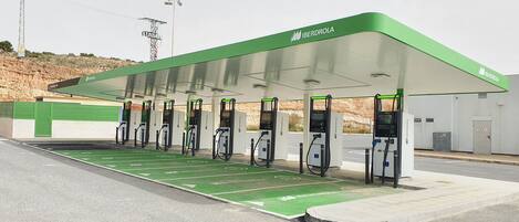 Una estación de carga para vehículos eléctricos con techo verde y varios puntos de carga, un área de espera cubierta y un espacio de estacionamiento pavimentado.