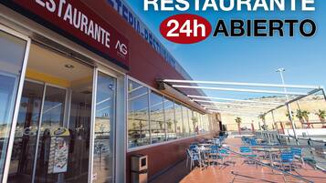 Un restaurante con letrero luminoso abierto las 24 horas, mesas al aire libre con sillas azules y vistas a un paisaje desértico.