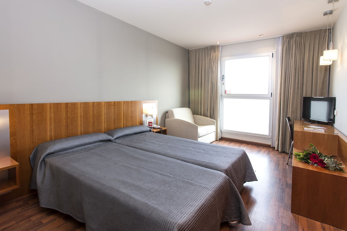 A hotel room with two beds, a desk with a computer, a chair, a TV, and a window with curtains.
