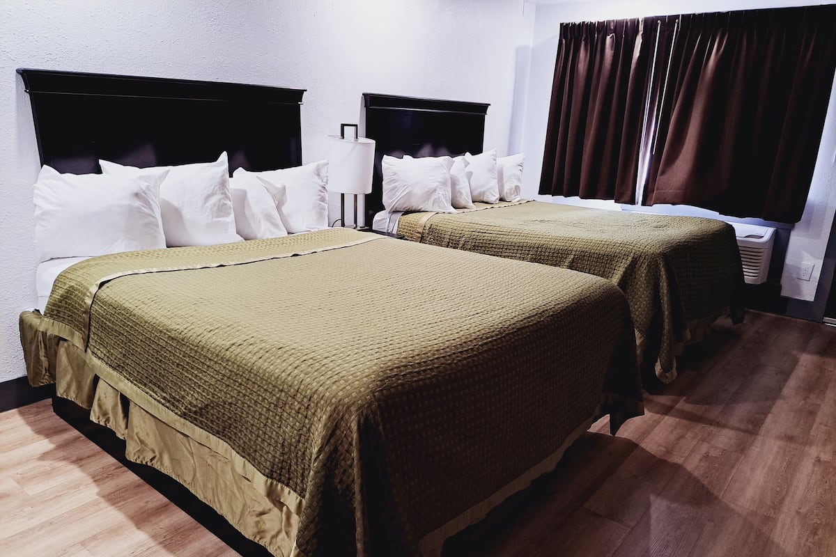 Une chambre d’hôtel avec deux lits, chacun doté d’une tête de lit et d’oreillers blancs. Une table de chevet avec une lampe se trouve entre les deux lits. La chambre a un plancher en bois et une fenêtre avec des rideaux.