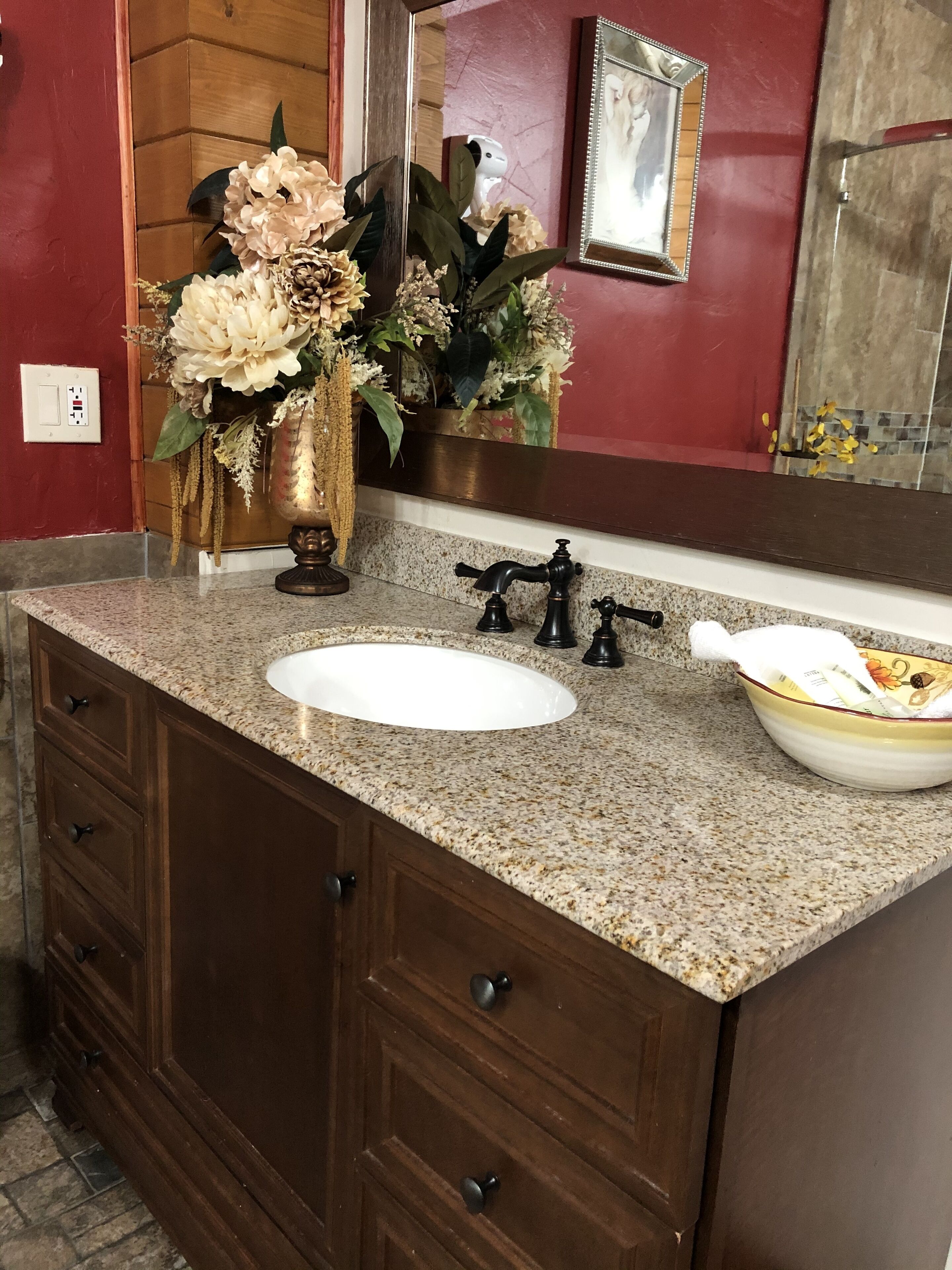 A bathroom with a granite countertop, a white sink, a mirror, a wooden vanity, and a vase with flowers.