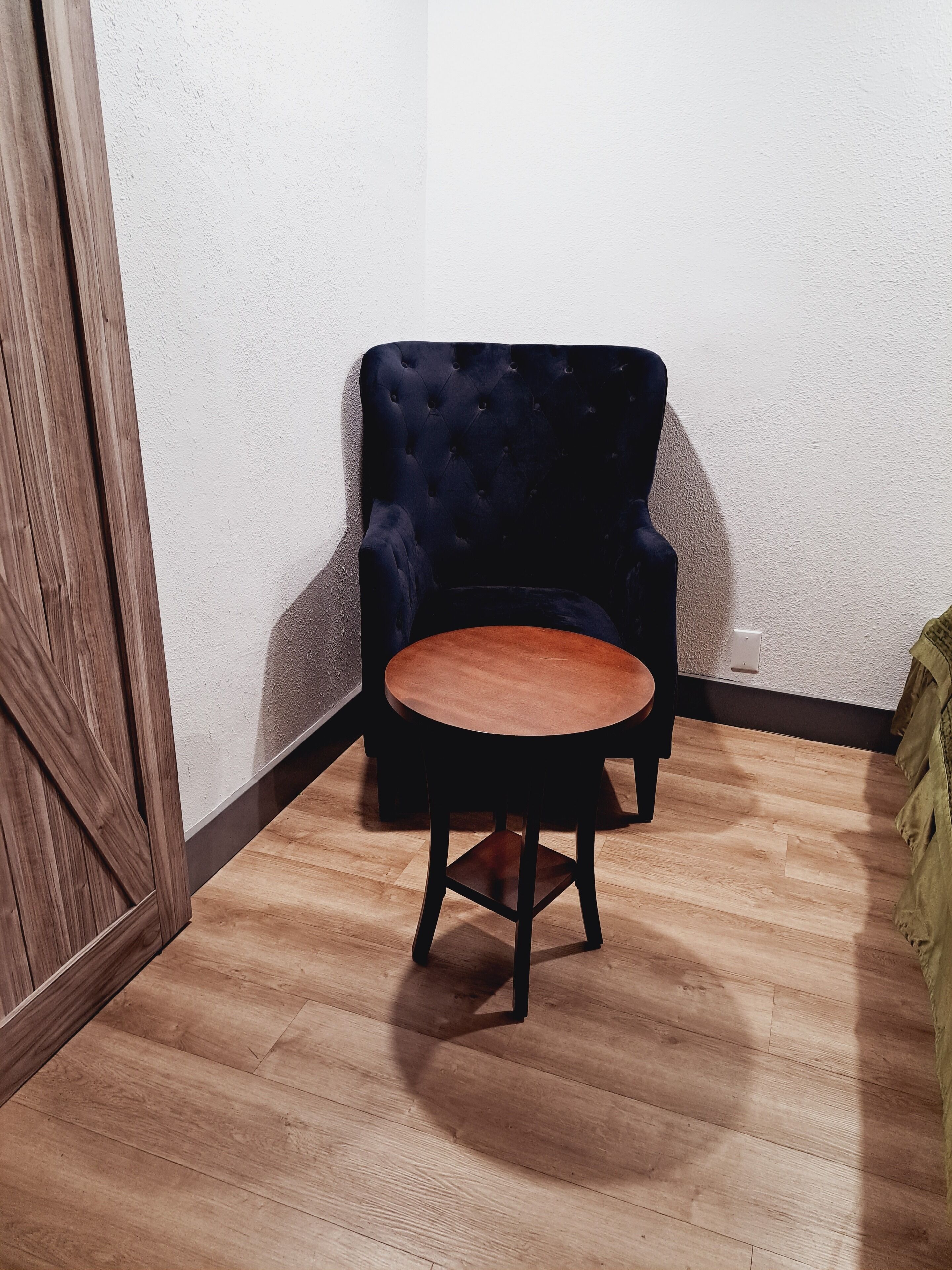 A corner room with a dark blue upholstered chair and a small round wooden table.