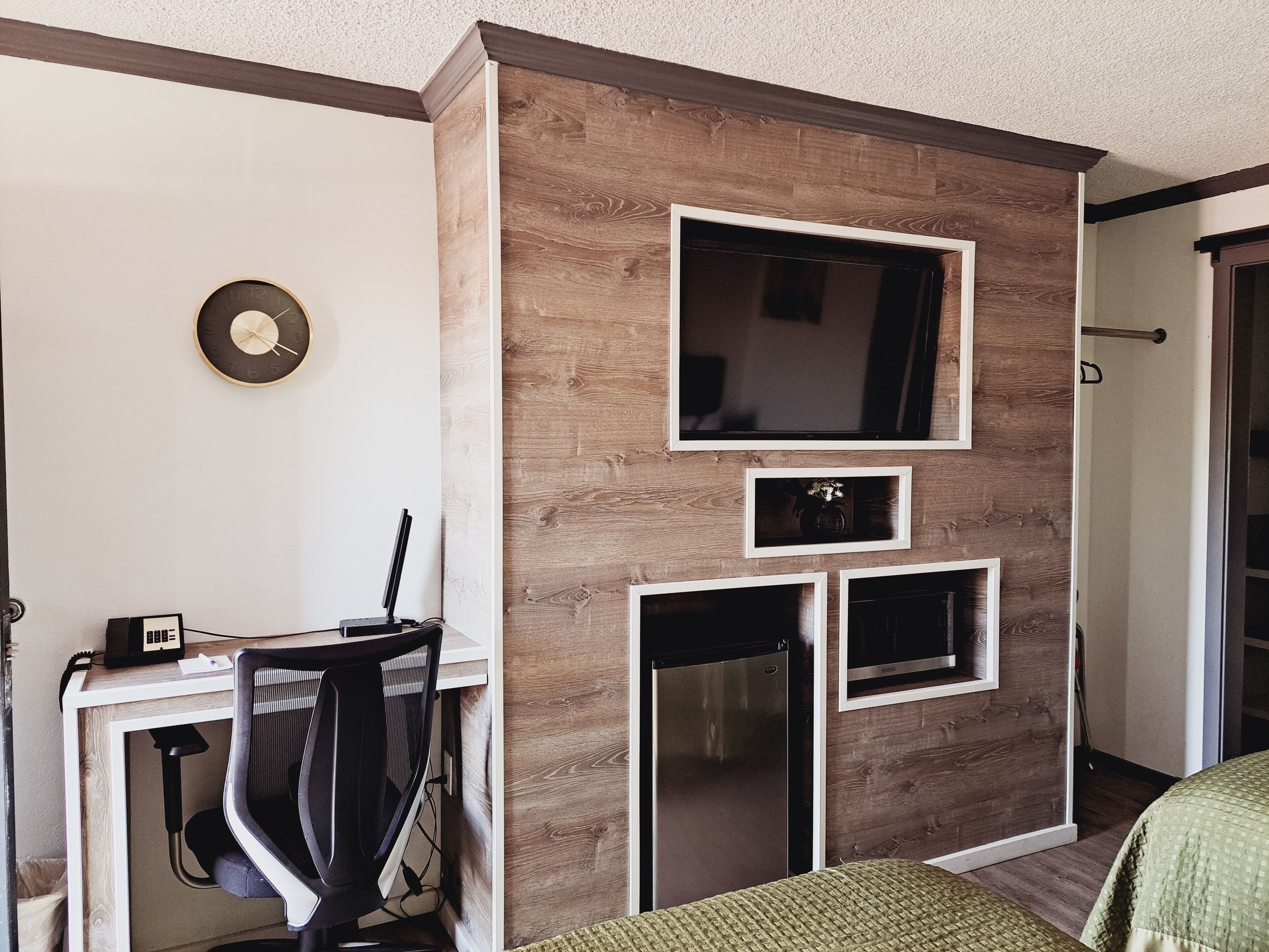 A compact hotel room with a wooden accent wall, a flat-screen TV, a small refrigerator, and a desk with a chair.