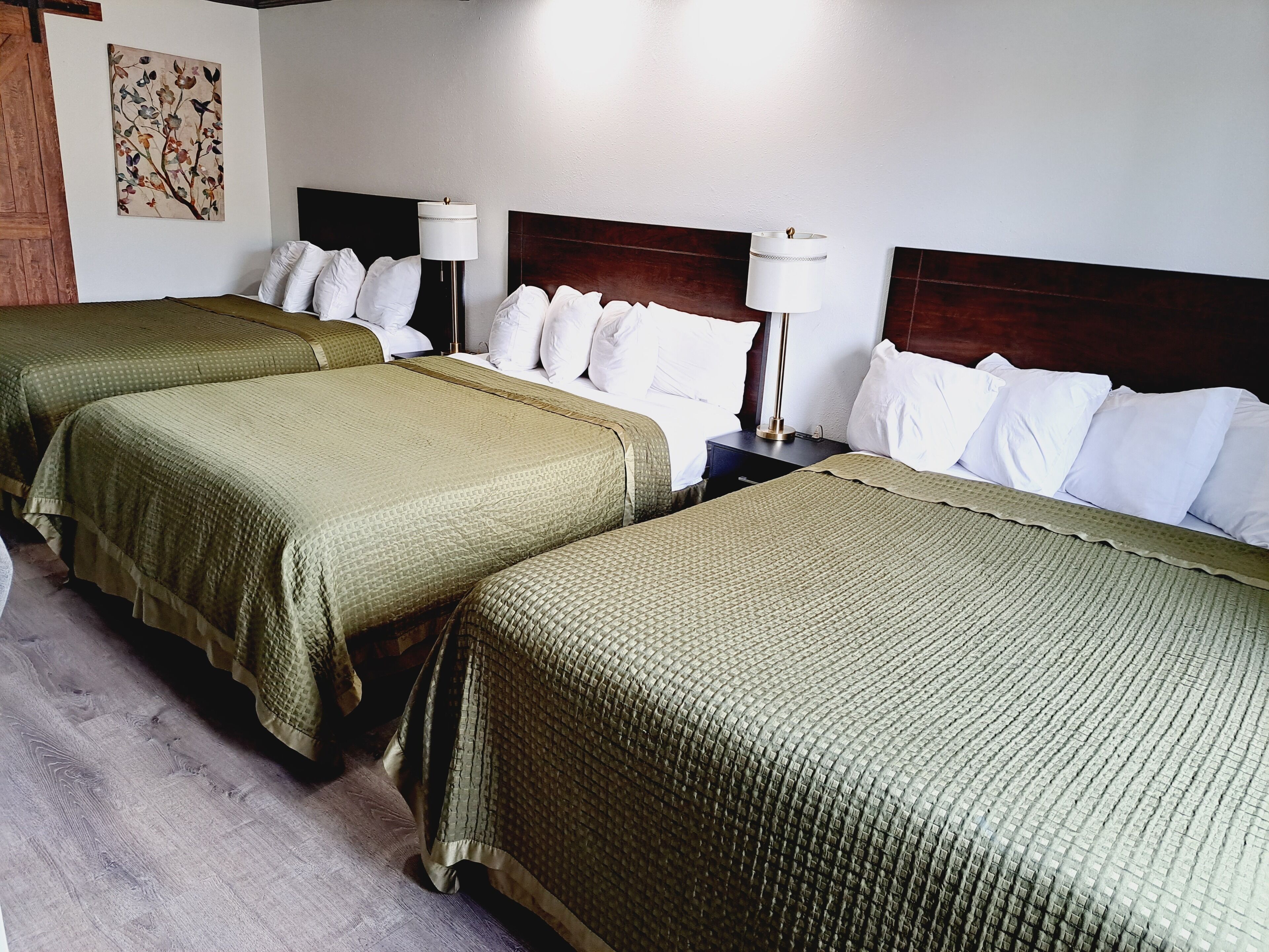 Habitación de hotel con tres camas, cada una con ropa de cama verde, almohadas blancas y cabeceros de madera. Hay una mesita de noche con una lámpara y una pequeña mesa también con una lámpara.