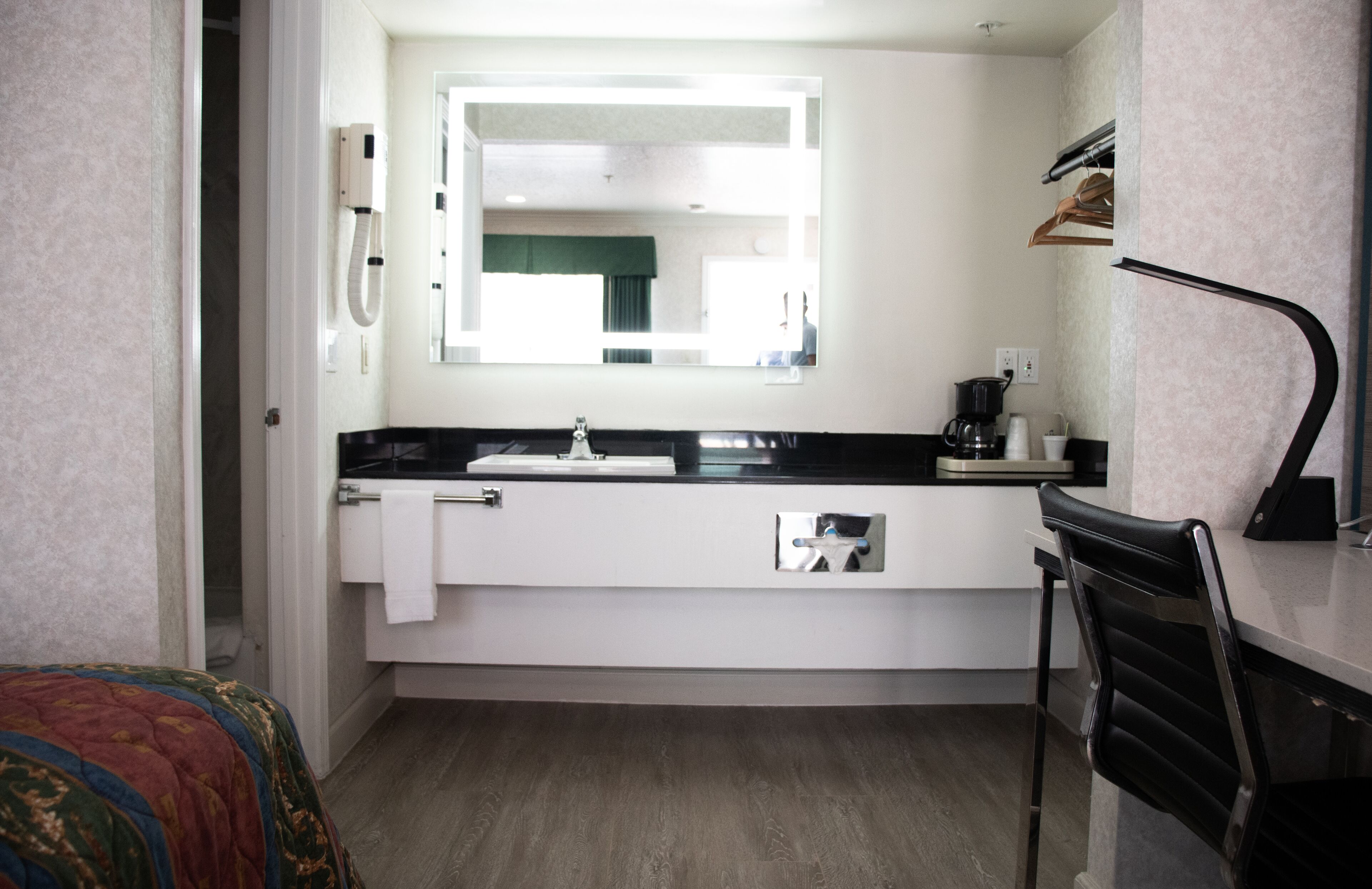 A compact hotel room with a white sink, a mirror above it, a black chair, and a bed with a colorful blanket.